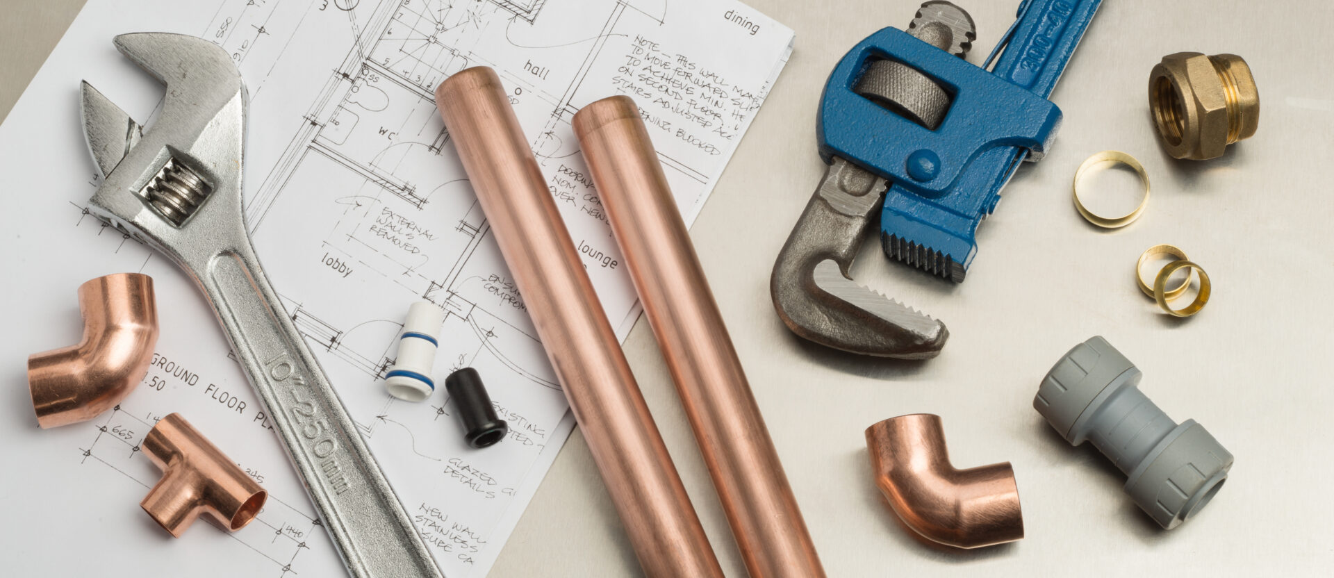 Plumbers tools and plumbing materials including copper pipe, elbow joint, wrench and spanner on top of architects house plans. MidCity Plumbers